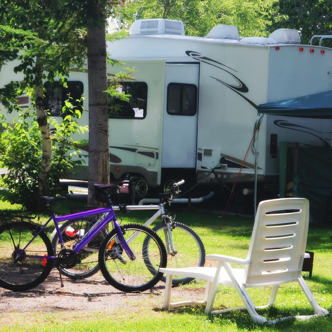 Camping scene with two bicycles, a lounge chair, and a motorhome among trees.
